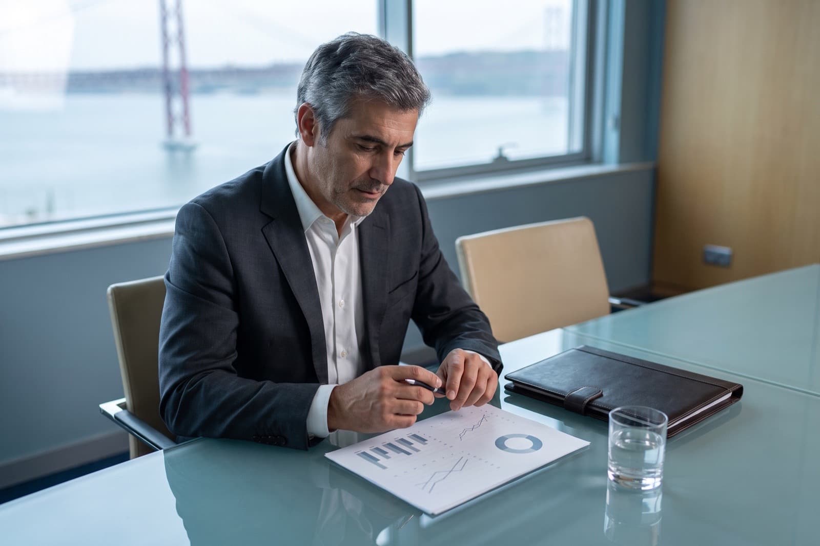 Fund manager in a charcoal suit reviewing a printed portfolio summary at a glass conference table with the Tagus river behind